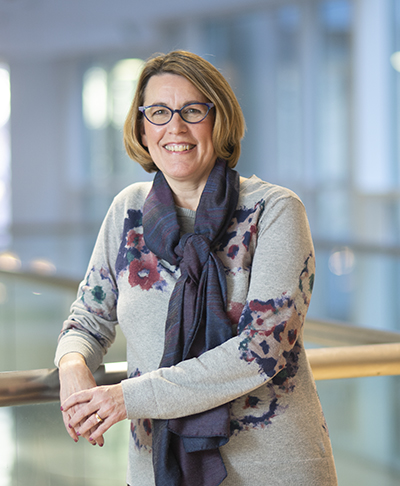 White woman wearing a sweater and scarf leans against banister and smiles at camera