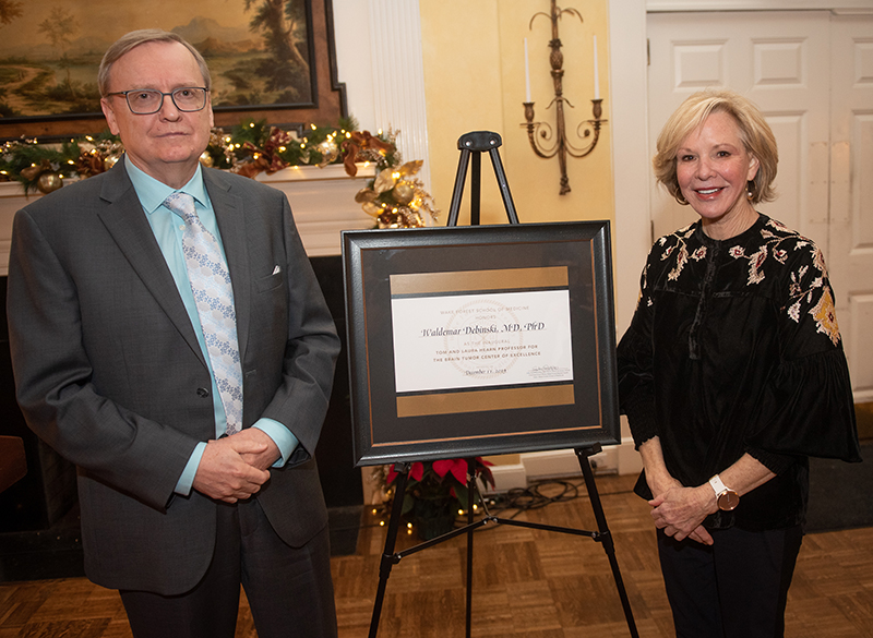 An older man with glasses and a blonde woman stand on either side of an easel