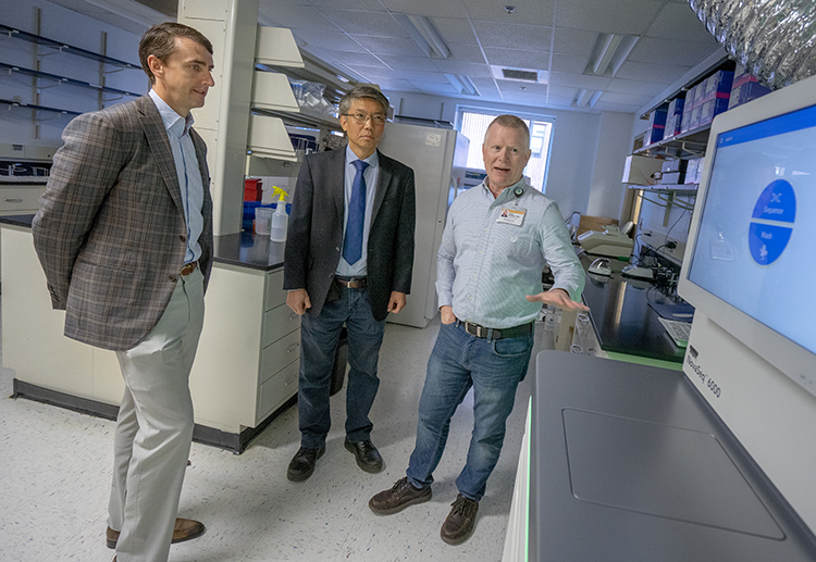 Three men stand in a lab and look at a large monitor