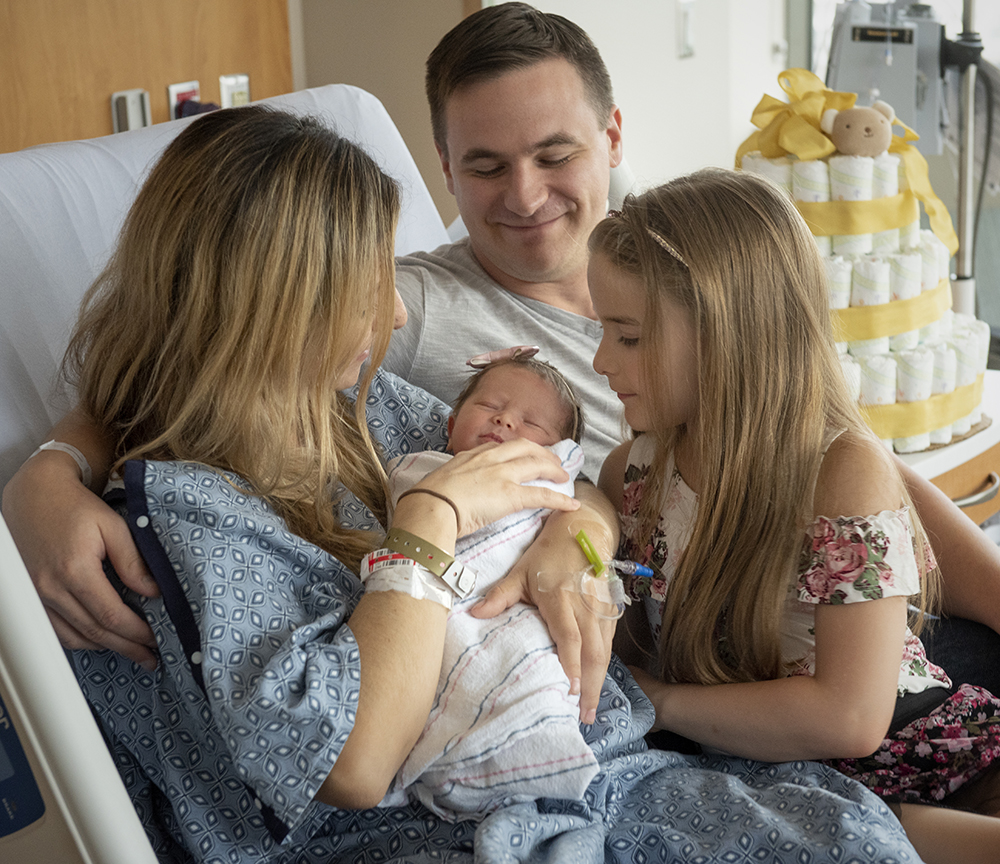 A mother, father and big sister look at a newborn girl as they sit on a hospital bed