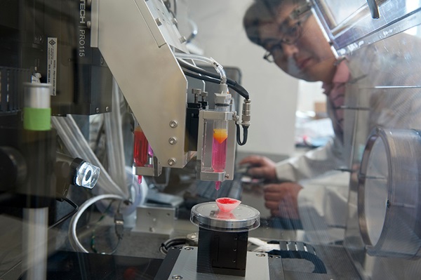A male scientist in a white coat looks at equipment in the regenerative laboratory as it drips red material into a mold