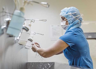 Stock image of heath care provider scrubbing up at a handwashing station and wearing blue scrubs, a hair net and face mask