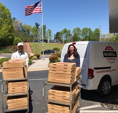 A man and woman stand behind carts full of pizza boxes, with a flag waving in the background
