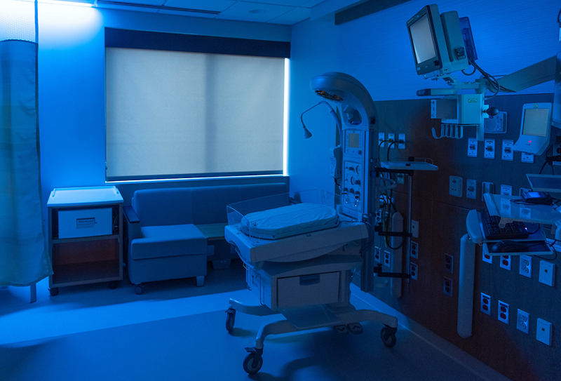 An empty NICU room with isolette, monitors and couch is bathed in blue light