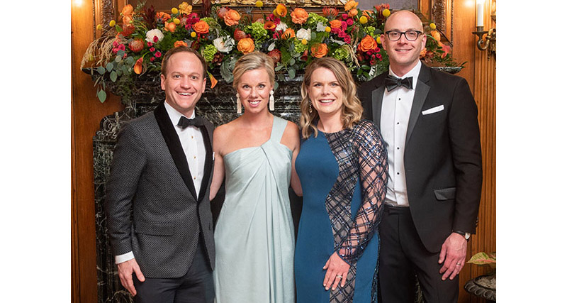 Two men and two women, all dressed in formal wear, stand in front of a portrait and floral arrangement