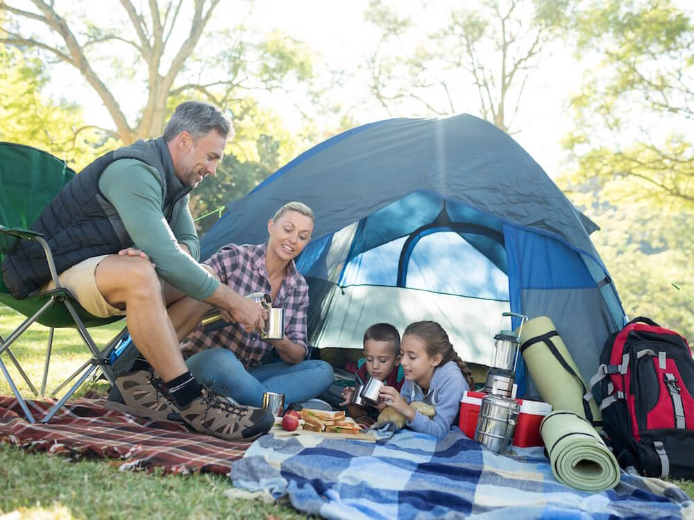 A family with two kids gathers together outside of a tent and next to camping equipment.