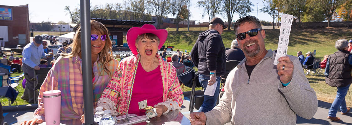 Two women in pink-patterned shirts, hats and glasses and one man in sunglasses and a white shirt smile while sitting at a table.