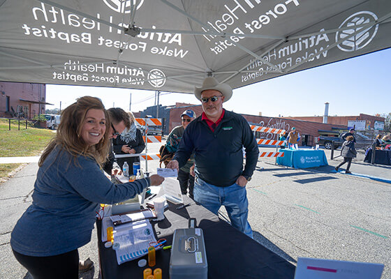 A man in a blue shirt and jeans and a woman in a blue shirt smile next to a merchandise display under an awning.