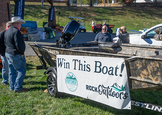 A group of men stand next to a boat that has a white banner saying, "Win this boat!" on it.