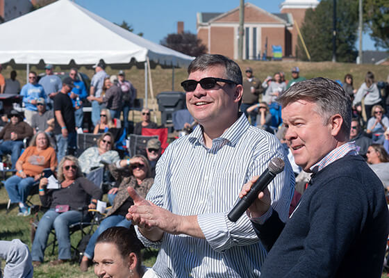 Two men in collared shirts making announcements on a microphone to a crowd.