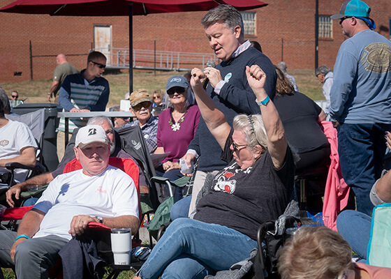 A woman in a gray t-shirt raising her arms in celebration while sitting in a small crowd.