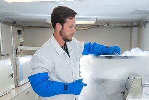 A man with dark hair wearing a white lab coat and blue gloves opening a container emitting steam.