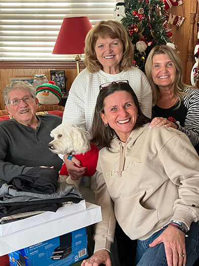 A group of family members in sweaters smile and stand together with their dog for a photo in front of a Christmas Tree.