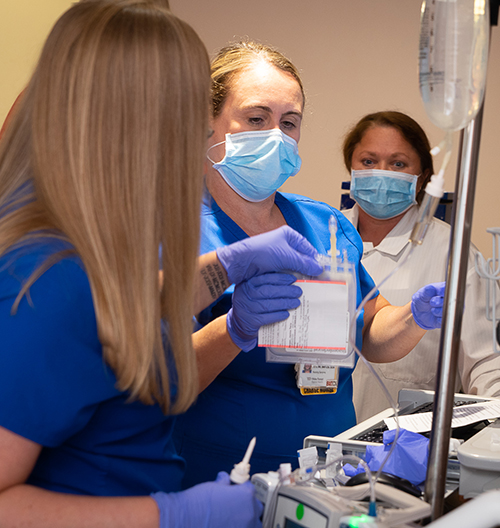 Two women in masks and blue scrubs look at empty fluid bag as woman in white coat watches