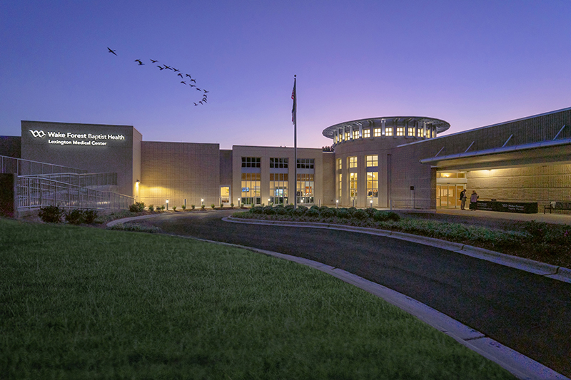 A large cream-colored building with circular entry glows with lights against a purple-blue evening sky