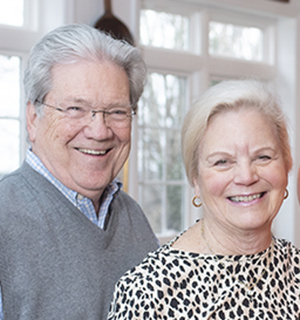 An older man and woman stand in front of some windows and smile at the camera
