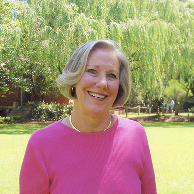 Blonde woman in bright pink shirt smiles at camera with trees, people and a brick building in the background