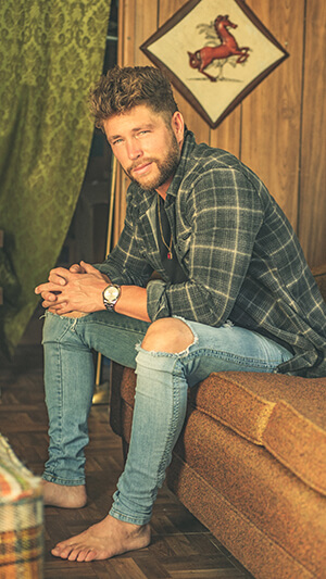 A young man wearing a plaid shirt and jeans sitting on a bench.