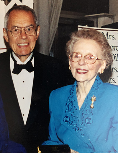 An older man and woman smile at the camera. He is wearing a tuxedo and she is in a turquoise formal dress
