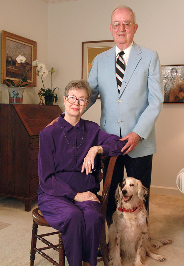An older woman wearing purple pants and blouse sits in a chair in a living room, and an older man wearing a light blue jacket and striped tie stands beside her with one hand on her elbow. A dog sits at their feet.