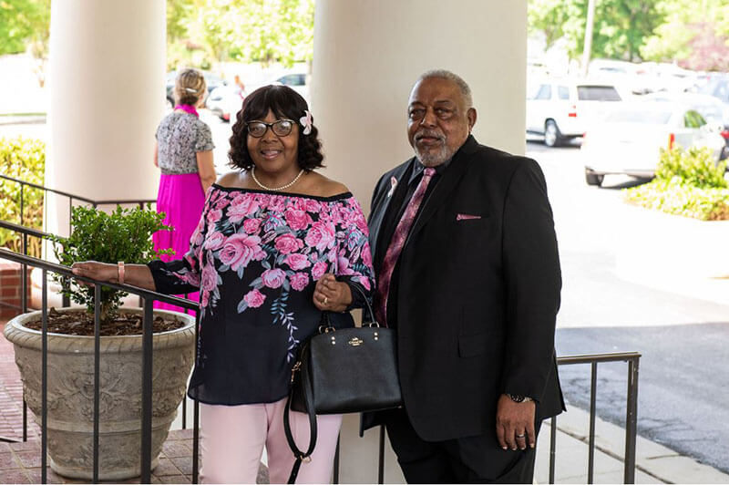 Ercell and Linda Tate outside of the Pink Ribbon Luncheon wearing formal attire standing by a staircase railing.