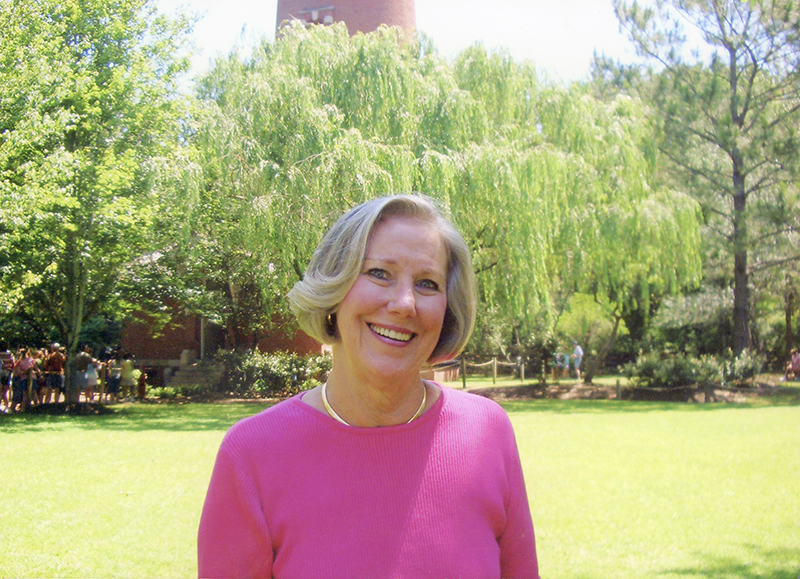 Blonde woman in bright pink shirt smiles at camera with trees, people and a brick building in the background