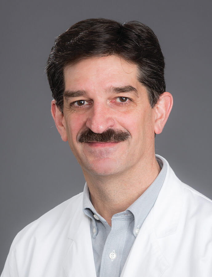 A man with dark hair wearing a lab coat and smiling at the camera.