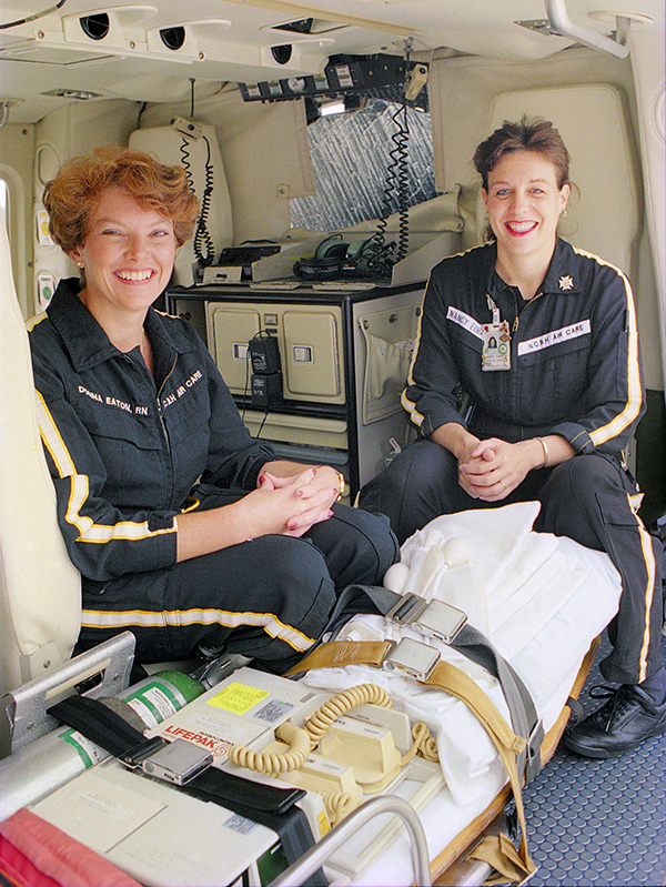 Two women in flight suits sit in the AirCare helicopter behind a gurney