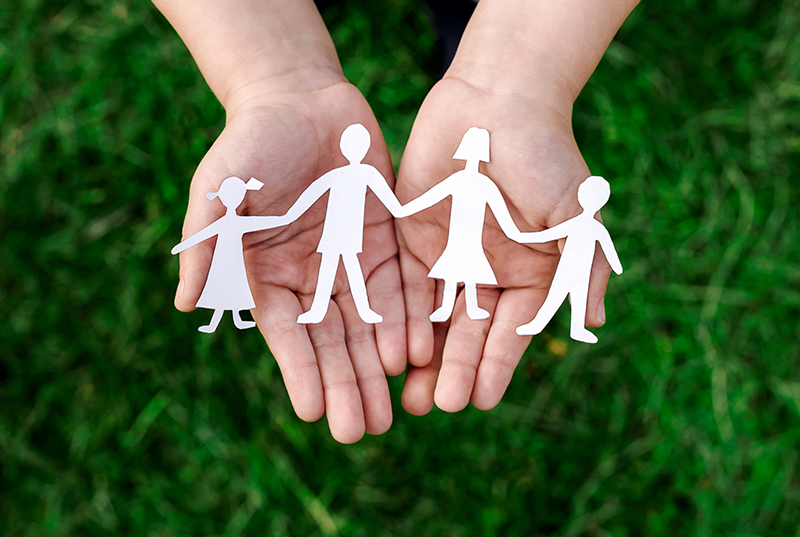 A pair of hands, palms up, holds a string of four white paper-doll cutouts over a background of green grass