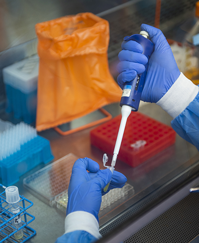 Hands in blue gloves and blue PPE sleeves hold a pipette and test tube, with an orange bag in the background
