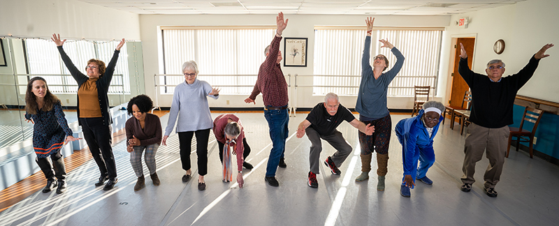 A group of older men and women stand in a line in a dance studio in various poses
