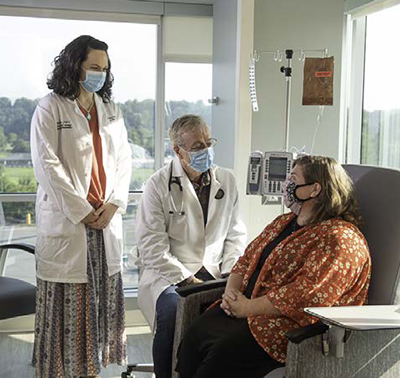 A standing woman and a seated man, both wearing face masks and white coats, talk to a woman sitting in a chair