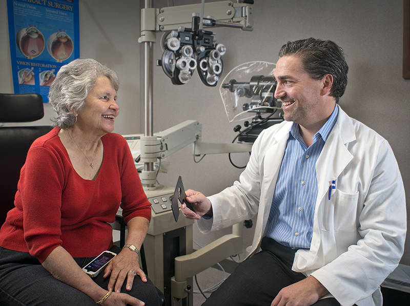 An older woman with gray hair and wearing a red top sits and smiles at a man in a white coat sitting in front of eye exam equipment