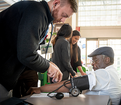 An older African American man wearing a hat sits in a chair and has his blood pressure taken by a younger white man