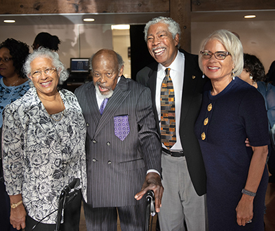 Two older African American couples stand together in a crowded room and smile at the camera