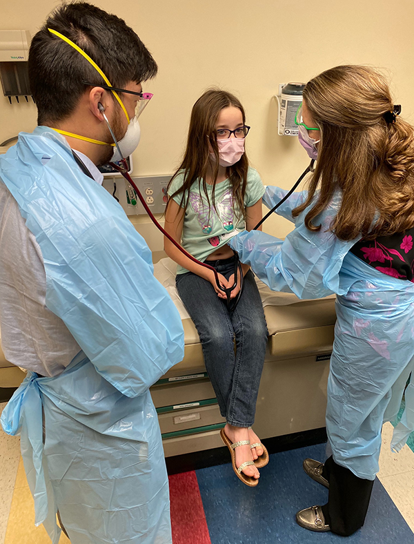 A girl with long brown hair and wearing jeans and a face mask sits on an exam table as a man and woman wearing blue gowns and face masks look at her