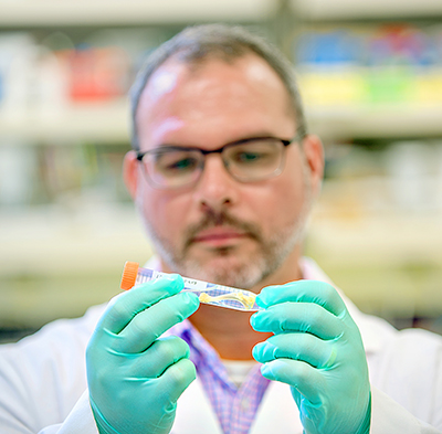 A man in glasses, gloves and a white coat looks at a test tube he is holding