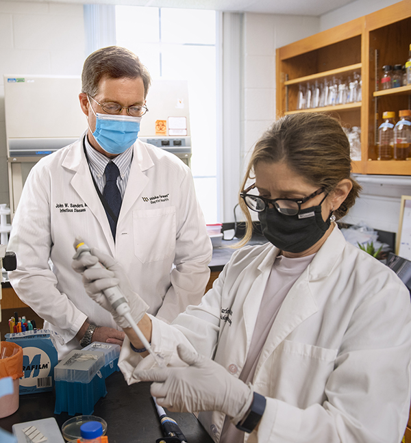 A man in white coat and facial mask watches a woman in a white coat and face mask use a pipette to add liquid to a test tube
