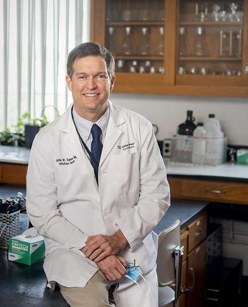 A man in a white coat and tie leans against a lab table and smiles at the camera