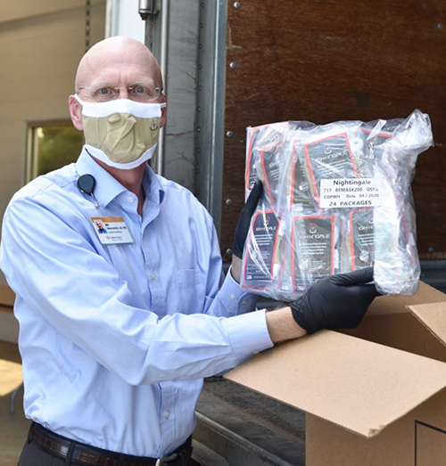 A bald man with glasses and wearing a blue shirt and tan face mask holds up a plastic-wrapped package of face masks from an open cardboard box