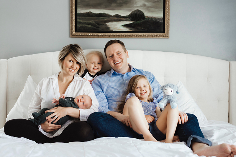 A man, woman, two young children and a baby sit on a bed with a white comforter and white upholstered headboard. They are close together and smiling at the camera