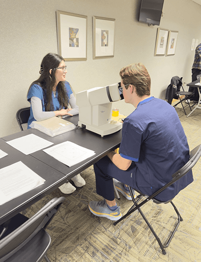 A young woman in blue scrubs administering a vision test to a man in dark blue scrubs.