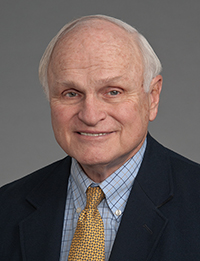 Older white man with white hair in dark jacket and yellow tie