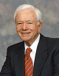 Older white man with full head of white hair in dark jacket and red striped tie