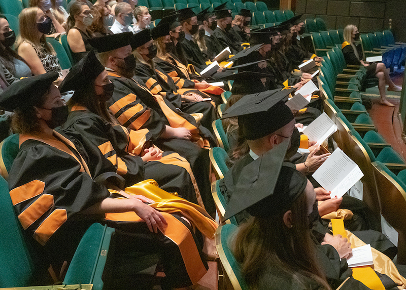 Two rows of men and women in caps and black graduation robes with gold stripes on the arms sit in green velvet seats. All are wearing black face masks. 