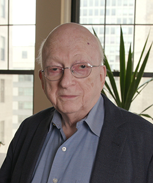 An elderly man wearing glasses, a blue button-down shirt and navy jacket poses for the camera in front of large windows overlooking a city