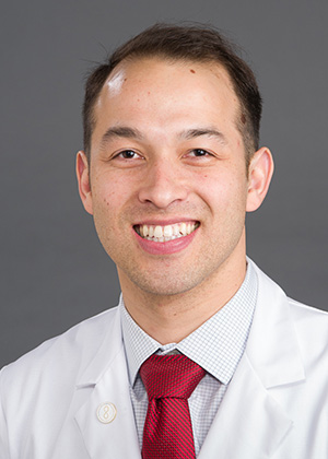 A dark-haired young man wearing a white coat and red tie smiles at the camera