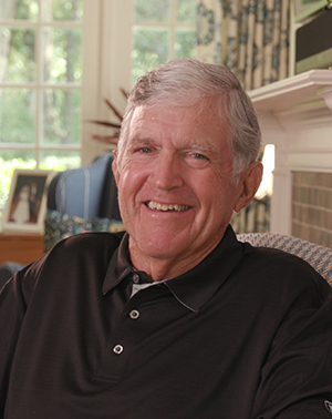 A man with white hair and wearing a navy golf shirt smiles as he sits in a living room