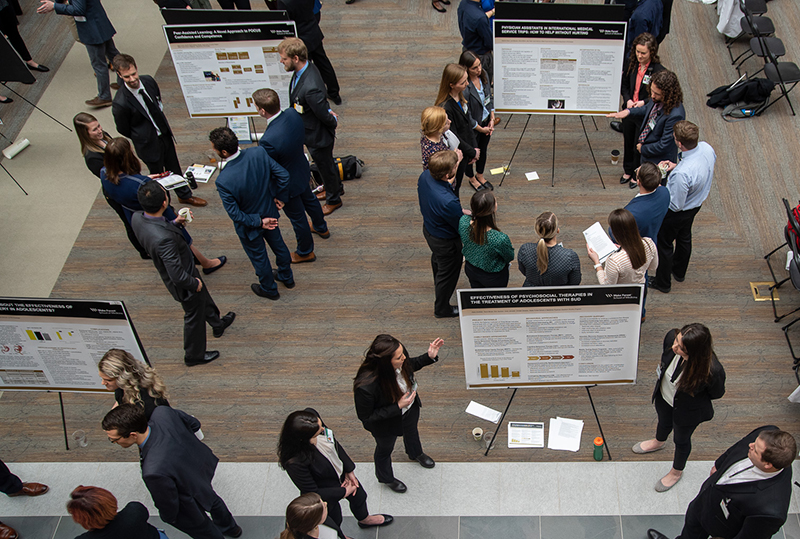 Overhead shot of men and women standing around research posters on easels, talking and listening