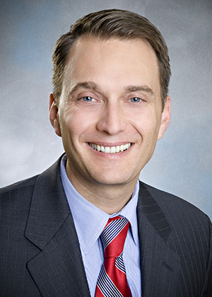 A dark-haired man wearing a blue button-down shirt, navy jacket and red and gray striped tie smiles in front of a blue background